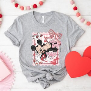 May include: A gray t-shirt featuring a graphic of Mickey and Minnie Mouse embracing, set within a pink postage stamp design. The design includes hearts, a bow, and a love letter. The shirt is displayed on a white wooden surface with Valentine's Day decorations.