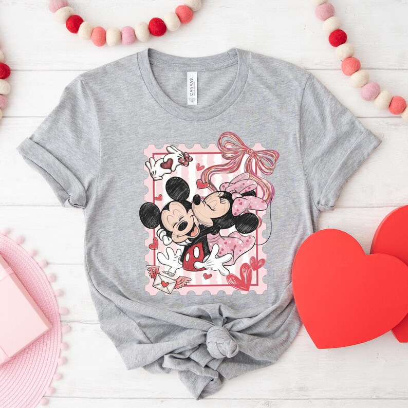 May include: A gray t-shirt featuring a graphic of Mickey and Minnie Mouse embracing, set within a pink postage stamp design. The design includes hearts, a bow, and a love letter. The shirt is displayed on a white wooden surface with Valentine's Day decorations.