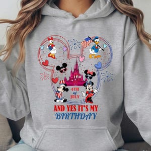 May include: A light gray hoodie featuring a colorful graphic of Disney characters celebrating the 4th of July. Mickey and Minnie Mouse, Donald and Daisy Duck are depicted with fireworks, hearts, and an American flag. Text reads "4th of July" and "And Yes It's My Birthday".
