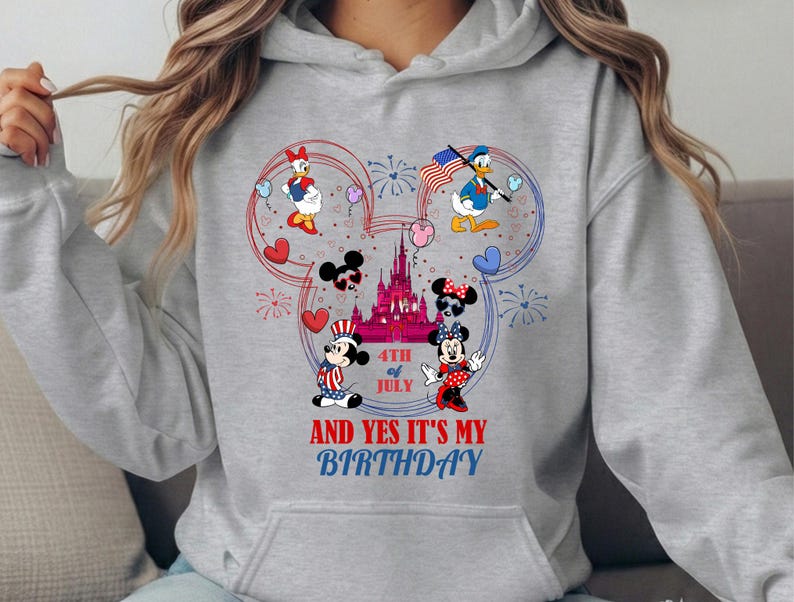 May include: A light gray hoodie featuring a colorful graphic of Disney characters celebrating the 4th of July. Mickey and Minnie Mouse, Donald and Daisy Duck are depicted with fireworks, hearts, and an American flag. Text reads "4th of July" and "And Yes It's My Birthday".