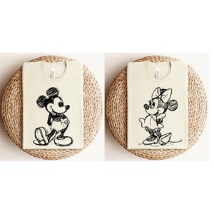 May include: Two cream-colored t-shirts with black and white cartoon images of Mickey Mouse and Minnie Mouse. The images are in a sketch style.