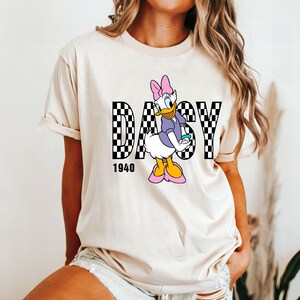May include: A white t-shirt with a black and white checkered graphic of the word "Daisy" with a cartoon image of Daisy Duck in the middle. The year "1940" is printed below the graphic.