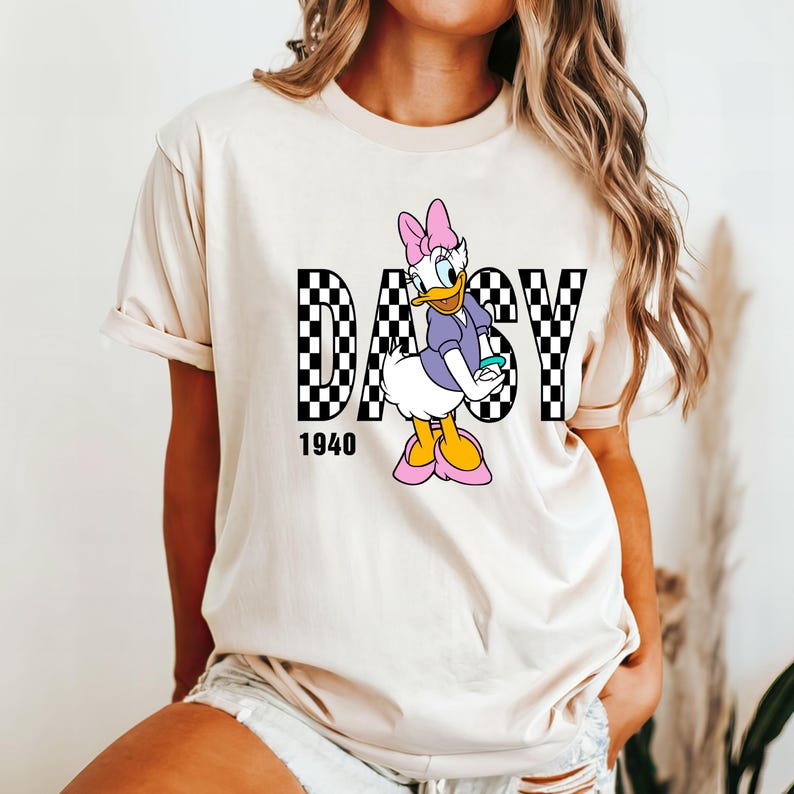 May include: A white t-shirt with a black and white checkered graphic of the word "Daisy" with a cartoon image of Daisy Duck in the middle. The year "1940" is printed below the graphic.