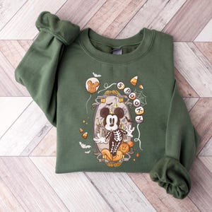 May include: Green sweatshirt featuring a Halloween-themed graphic. The design includes a skeleton Mickey Mouse, a haunted house, bats, and the words "Spooky" and "Mickey Mouse". The sweatshirt has long sleeves with ribbed cuffs and a crew neck.