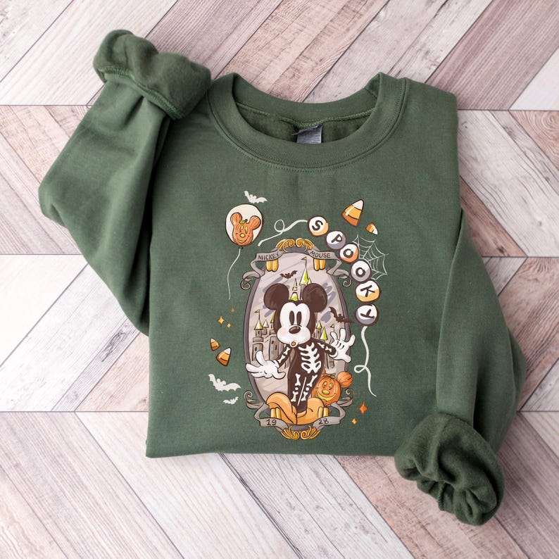 May include: Green sweatshirt featuring a Halloween-themed graphic. The design includes a skeleton Mickey Mouse, a haunted house, bats, and the words "Spooky" and "Mickey Mouse". The sweatshirt has long sleeves with ribbed cuffs and a crew neck.