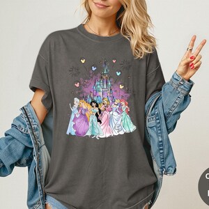 K&ouml;nnte beinhalten: Dunkelgraues T-Shirt mit einem farbenfrohen Aufdruck von Disney-Prinzessinnen vor einem Schloss, mit Feuerwerk und herzf&ouml;rmigen Ballons. Das T-Shirt ist mit "Comfort Colors Pepper" gekennzeichnet.