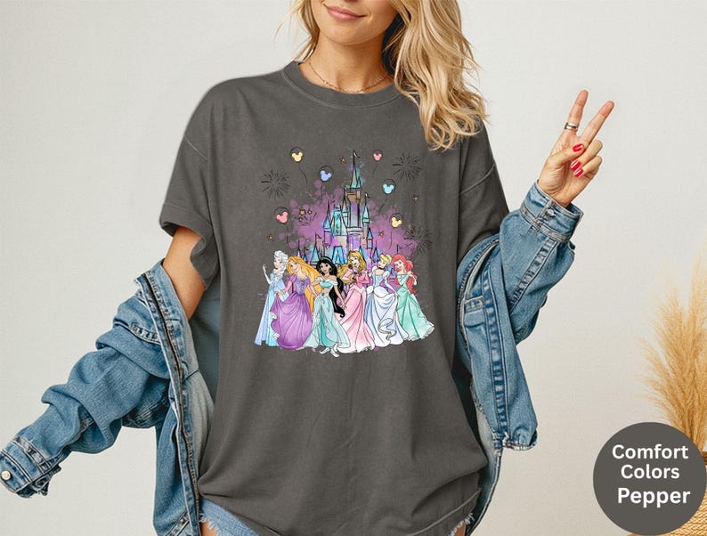 K&ouml;nnte beinhalten: Dunkelgraues T-Shirt mit einem farbenfrohen Aufdruck von Disney-Prinzessinnen vor einem Schloss, mit Feuerwerk und herzf&ouml;rmigen Ballons. Das T-Shirt ist mit "Comfort Colors Pepper" gekennzeichnet.