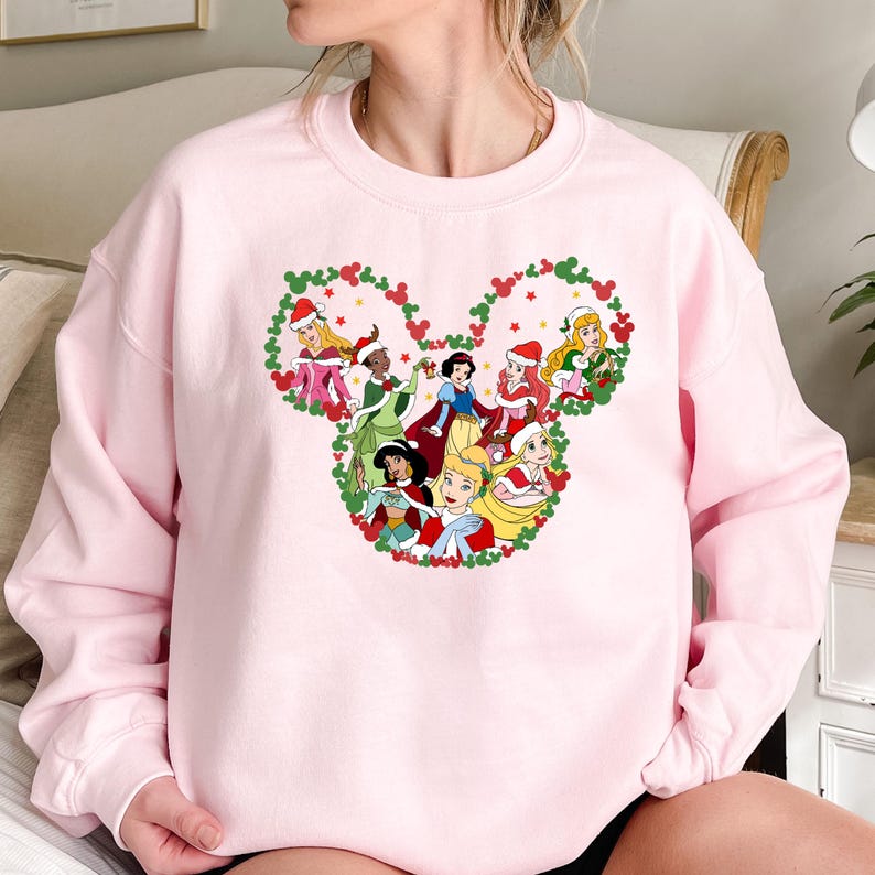 Pu&ograve; includere: Una felpa rosa chiaro con un design a forma di testa di Topolino riempita di principesse dei cartoni animati in abiti festivi. Le principesse sono incorniciate da una ghirlanda verde con bacche rosse e stelle. La felpa &egrave; girocollo.