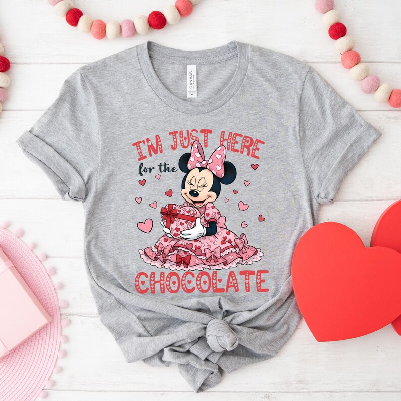 May include: A gray t-shirt featuring a graphic of Minnie Mouse in a pink dress holding a heart-shaped gift box. The text "I'M JUST HERE for the CHOCOLATE" is printed in red. The design includes pink hearts.