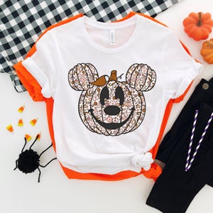 May include: White t-shirt with a floral pumpkin Mickey Mouse design. The pumpkin has a brown stem and a smiling face. The shirt is a popular Halloween design.