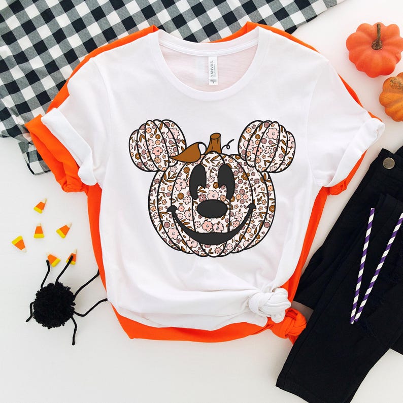 May include: White t-shirt with a floral pumpkin Mickey Mouse design. The pumpkin has a brown stem and a smiling face. The shirt is a popular Halloween design.