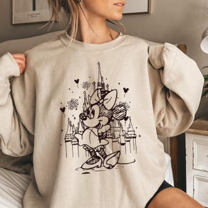 May include: A beige sweatshirt with a black and white graphic print of Mickey Mouse in front of a castle. The graphic also includes fireworks and hearts.