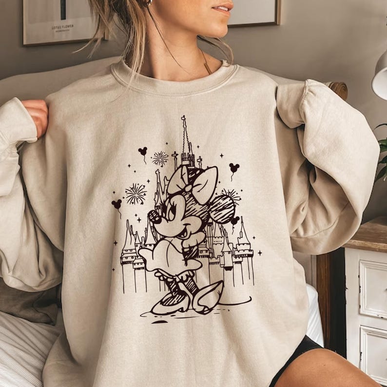 May include: A beige sweatshirt with a black and white graphic print of Mickey Mouse in front of a castle. The graphic also includes fireworks and hearts.