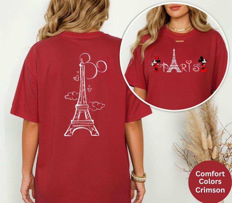 May include: Crimson red t-shirt featuring a white Eiffel Tower design with Mickey and Minnie Mouse characters. The shirt has a relaxed fit and the word "PARIS" is printed in white. The back of the shirt also features an Eiffel Tower design.