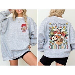May include: A heather gray sweatshirt featuring a Daisy Duck graphic on the front and a Christmas-themed design on the back. The back design includes Mickey Mouse, Minnie Mouse, Donald Duck, and other characters with the words "Very Merry Christmas".