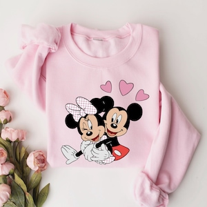 May include: Pink sweatshirt with a graphic of Mickey Mouse and Minnie Mouse hugging, surrounded by pink hearts.