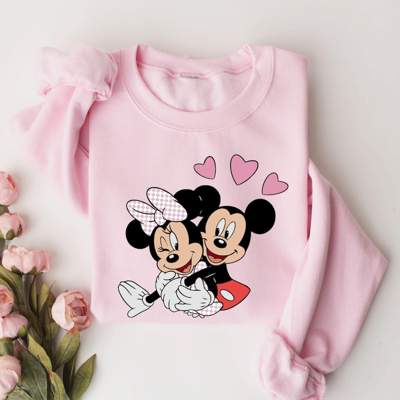 May include: Pink sweatshirt with a graphic of Mickey Mouse and Minnie Mouse hugging, surrounded by pink hearts.