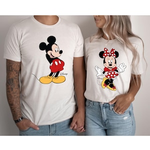 K&ouml;nnte beinhalten: Ein wei&szlig;es T-Shirt mit einem Cartoon-Bild von Mickey Mouse in roten Shorts und gelben Schuhen. Das Shirt hat den Schriftzug "Disney" aufgedruckt. Ein wei&szlig;es T-Shirt mit einem Cartoon-Bild von Minnie Mouse in einem roten, gepunkteten Kleid und roten Schuhen. Das Shirt hat den Schriftzug "Disney" aufgedruckt.