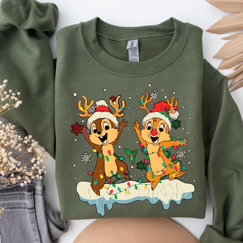 May include: Green Christmas sweatshirt featuring a cartoon image of two chipmunks wearing Santa hats and reindeer antlers, with Christmas lights wrapped around them. The chipmunks are standing on a snowy ground with a background of snowflakes.