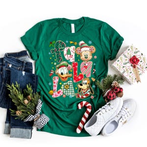 K&ouml;nnte beinhalten: Ein gr&uuml;nes T-Shirt mit dem Schriftzug "FA LA LA" in einem festlichen Design mit Mickey Mouse, Donald Duck und Pluto. Das Shirt wird mit blauen Jeans, wei&szlig;en Turnschuhen und einem verpackten Geschenk kombiniert.