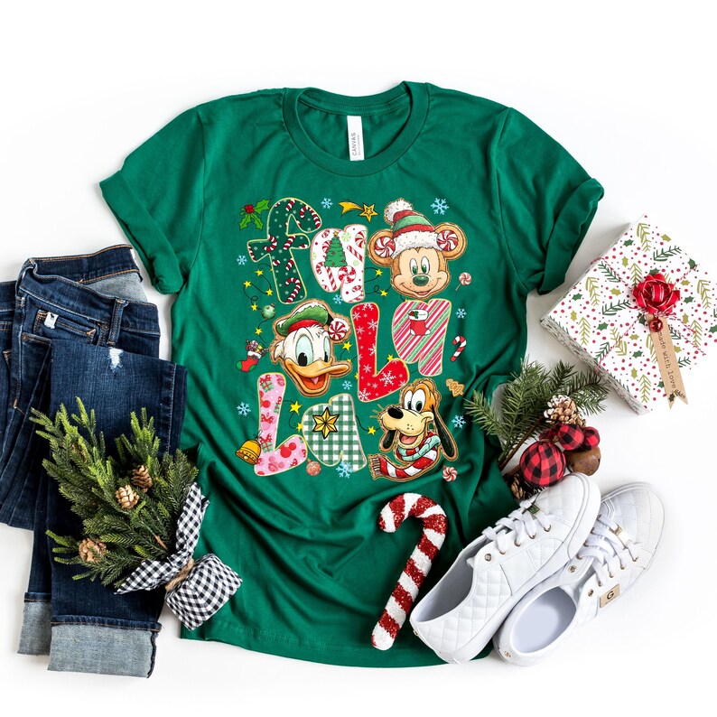 K&ouml;nnte beinhalten: Ein gr&uuml;nes T-Shirt mit dem Schriftzug "FA LA LA" in einem festlichen Design mit Mickey Mouse, Donald Duck und Pluto. Das Shirt wird mit blauen Jeans, wei&szlig;en Turnschuhen und einem verpackten Geschenk kombiniert.