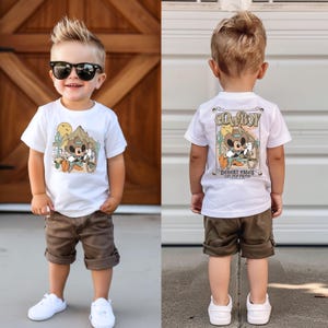 May include: A white t-shirt with a cartoon character wearing a cowboy hat and riding a horse. The text on the shirt says "Cowboy" and "Desert Miles on my Mind". The shirt is being worn by a young boy wearing brown shorts.