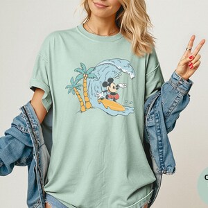 May include: A light green t-shirt featuring a graphic of Mickey Mouse surfing a wave. The design includes palm trees and a yellow surfboard. The shirt is a Comfort Colors brand.