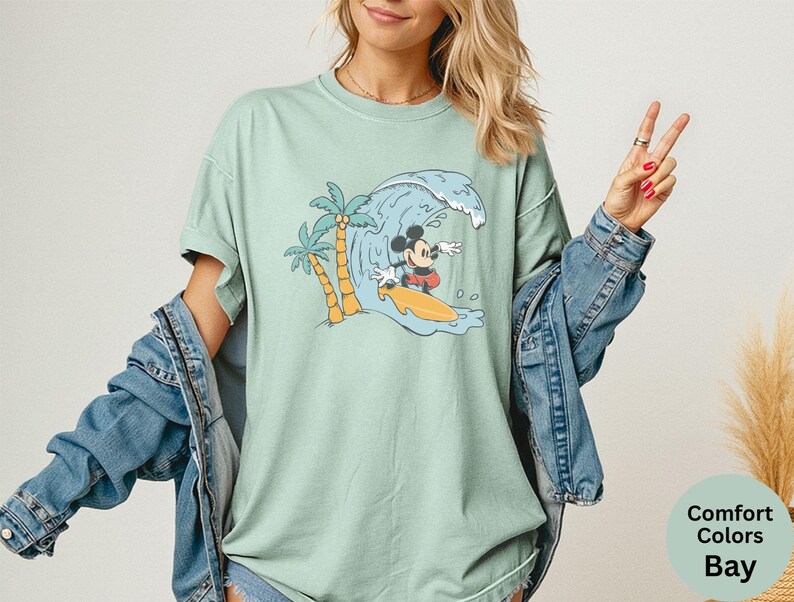 May include: A light green t-shirt featuring a graphic of Mickey Mouse surfing a wave. The design includes palm trees and a yellow surfboard. The shirt is a Comfort Colors brand.