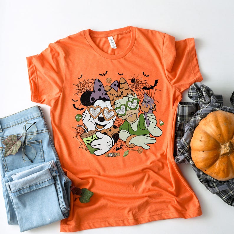 May include: Orange t-shirt with a graphic of Mickey Mouse and Daisy Duck wearing heart-shaped sunglasses and holding a spooky drink. The graphic is surrounded by bats, spiderwebs, and candy.