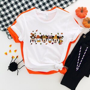 May include: White t-shirt with a colorful graphic featuring cartoon characters in fall-themed attire. The design includes images of Mickey Mouse, Minnie Mouse, Donald Duck, and Goofy. The shirt is paired with black pants and Halloween decorations.