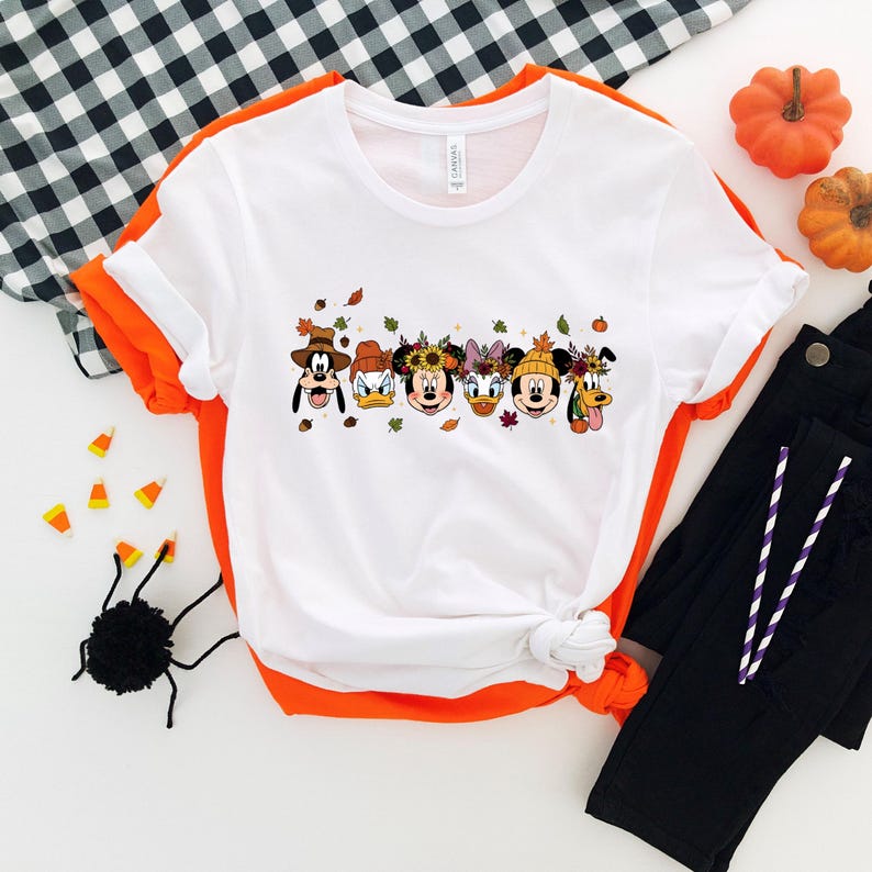 May include: White t-shirt with a colorful graphic featuring cartoon characters in fall-themed attire. The design includes images of Mickey Mouse, Minnie Mouse, Donald Duck, and Goofy. The shirt is paired with black pants and Halloween decorations.
