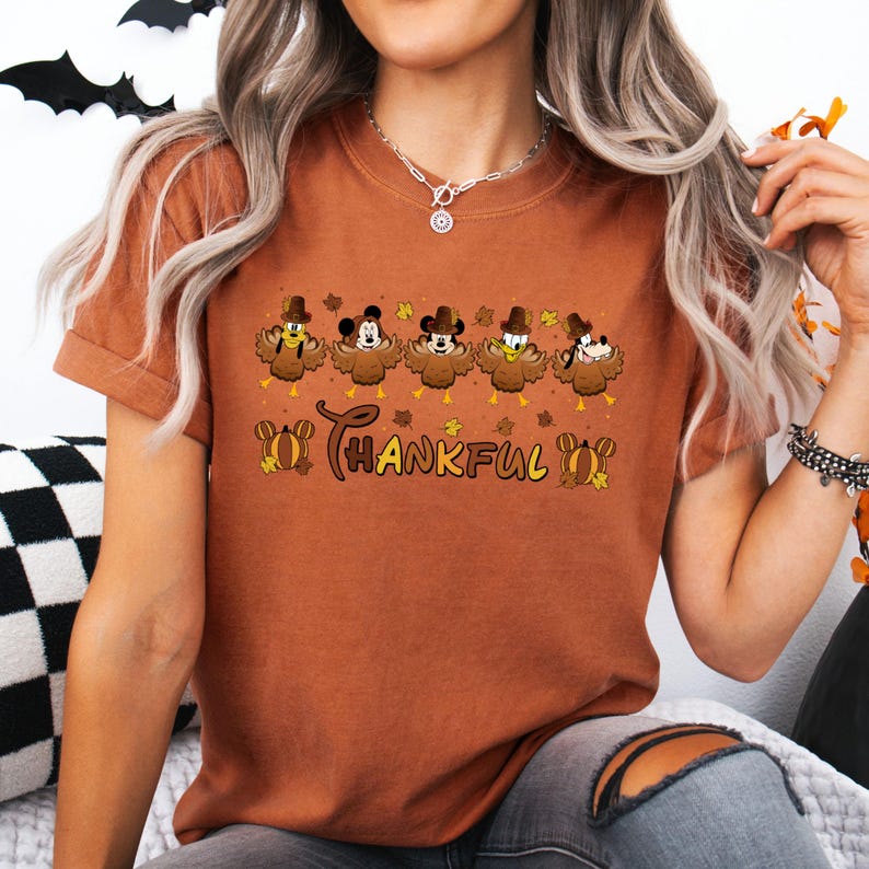 May include: A rust-colored t-shirt featuring a Thanksgiving design. The graphic includes cartoon turkey characters dressed as Mickey Mouse, Minnie Mouse, Donald Duck, and Goofy, with the word "THANKFUL" in a script font.
