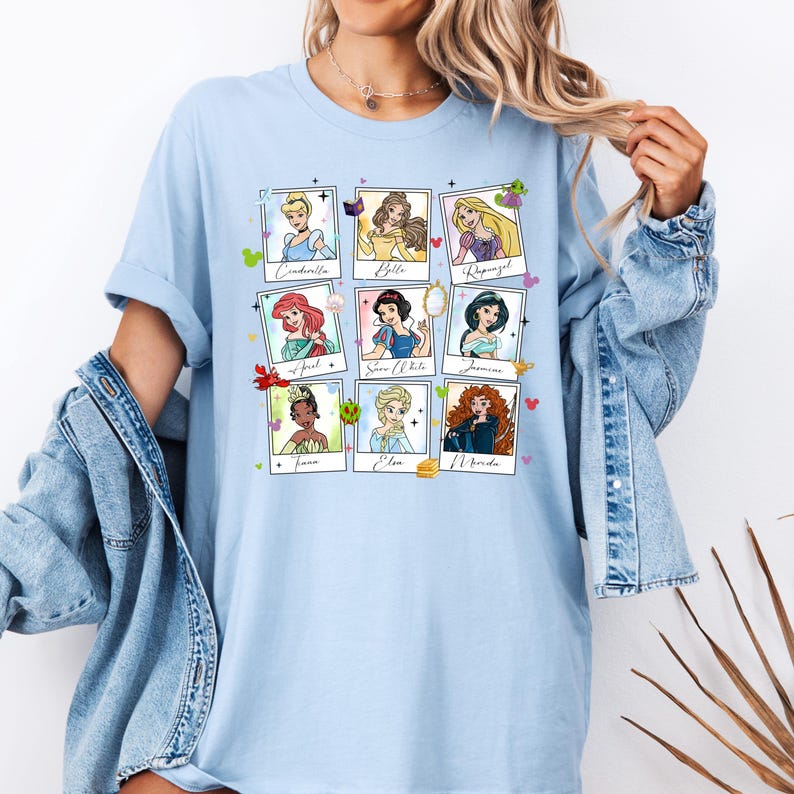 May include: Light blue t-shirt featuring a grid of nine Disney princess portraits in polaroid-style frames. Each frame includes the princess's name. The shirt is a casual, comfortable style.