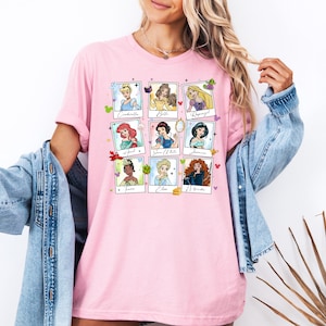 May include: Pink t-shirt featuring nine Disney princess portraits in a grid layout. Each portrait is framed with a white border and includes the princess's name. The shirt is a soft, casual style.