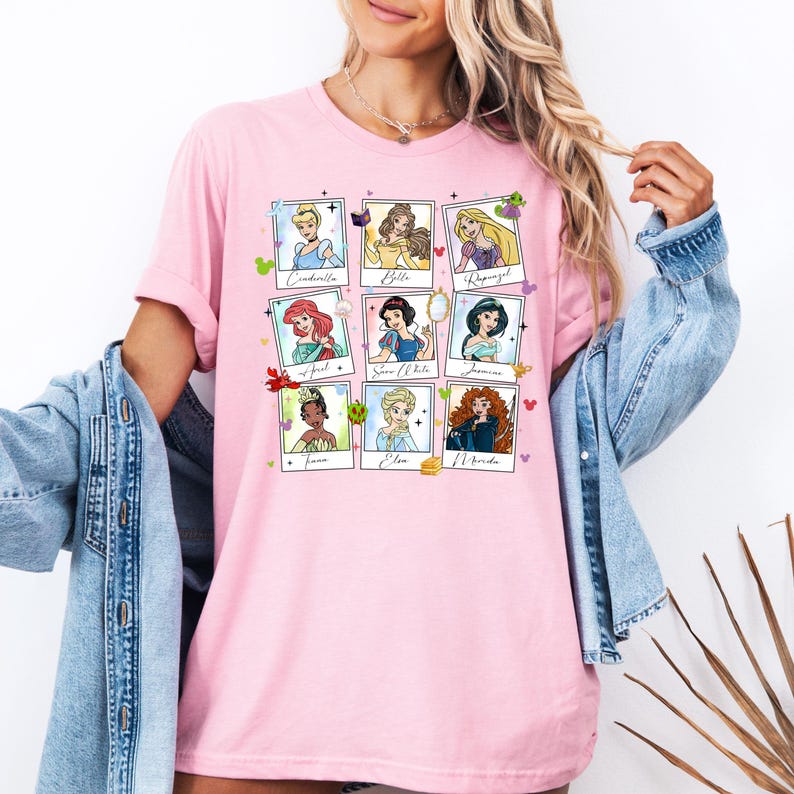 May include: Pink t-shirt featuring nine Disney princess portraits in a grid layout. Each portrait is framed with a white border and includes the princess's name. The shirt is a soft, casual style.