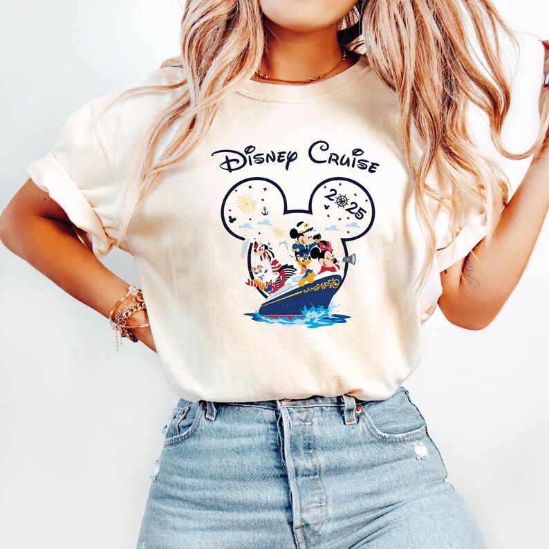 May include: Cream-colored t-shirt featuring a Disney Cruise design. The graphic includes Mickey Mouse, Minnie Mouse, and other characters on a cruise ship, with the words "Disney Cruise 2025" in a Mickey Mouse head outline. The shirt is a casual style.