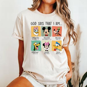 Puede incluir: Una camiseta beige con un gr&aacute;fico colorido con personajes de Disney y el texto "GOD SAYS THAT I AM...". Cada personaje est&aacute; emparejado con una afirmaci&oacute;n positiva y una referencia b&iacute;blica. La camiseta tiene mangas cortas.