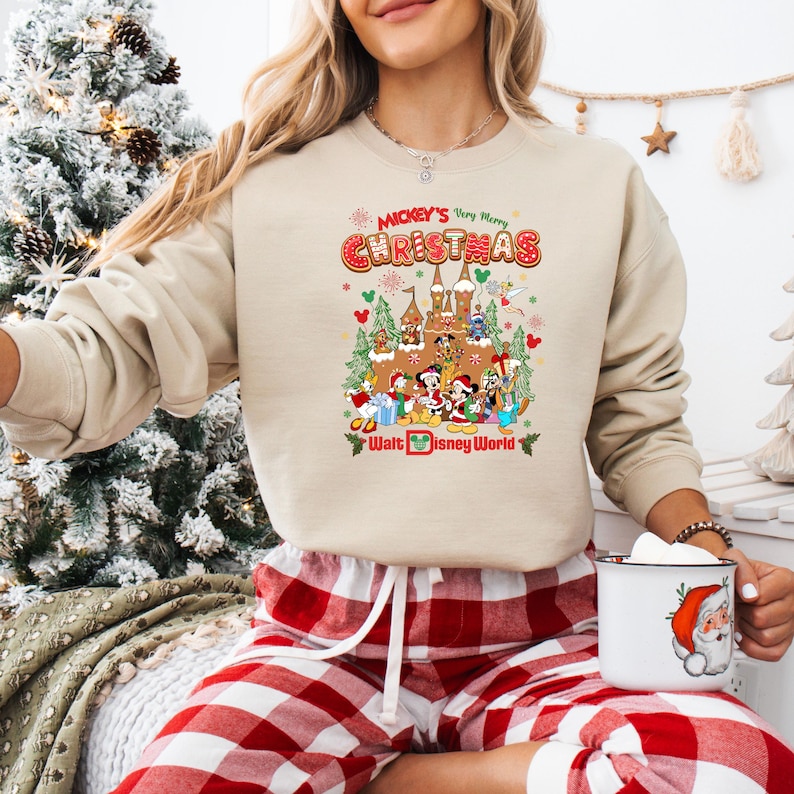 May include: Beige sweatshirt featuring a colorful graphic of Disney characters celebrating Christmas in front of a castle. The text "Mickey's Very Merry Christmas" and "Walt Disney World" are also displayed.