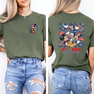 Puede incluir: Camiseta verde oliva con un dise&ntilde;o patri&oacute;tico de dibujos animados. La parte delantera tiene un personaje de dibujos animados con un sombrero patri&oacute;tico. La parte trasera muestra un grupo de personajes de dibujos animados con banderas y fuegos artificiales, con el texto "4th of July" y "Est. 1776".
