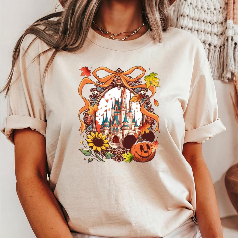 May include: Beige t-shirt featuring a colorful graphic of a castle inside an ornate frame with autumn leaves, a sunflower, and a jack-o'-lantern. The design includes Mickey Mouse ears and a bow. The shirt has short sleeves.