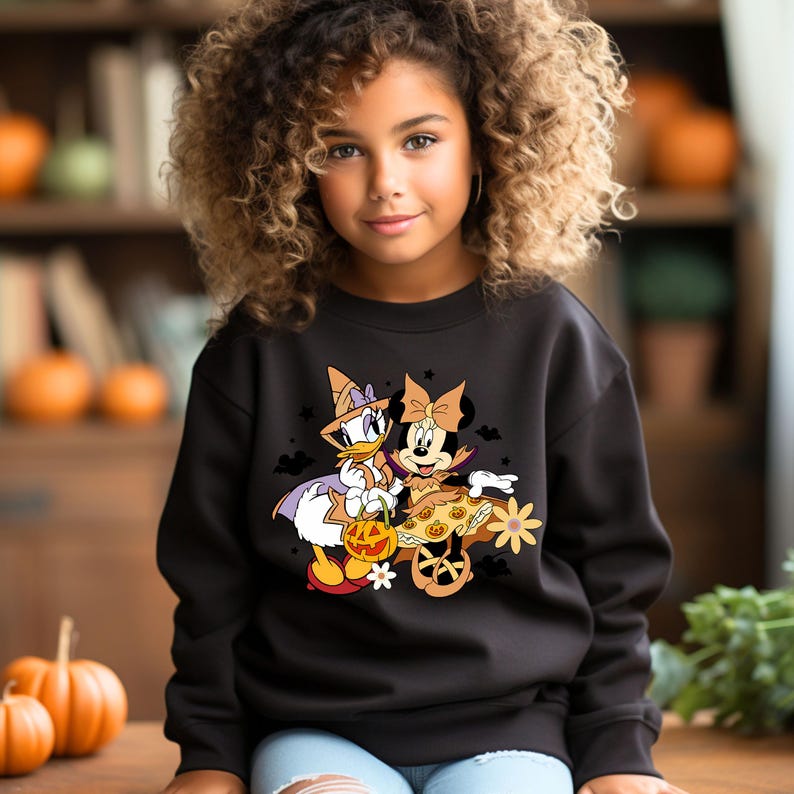 May include: Black sweatshirt with a graphic print of Minnie Mouse and Daisy Duck dressed in Halloween costumes. Minnie Mouse is wearing a vampire costume and Daisy Duck is wearing a witch costume. They are both holding a pumpkin.