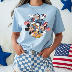 May include: Light blue t-shirt featuring a graphic of Mickey Mouse, Minnie Mouse, Donald Duck, Daisy Duck, Goofy, and Pluto holding American flags. The characters are wearing patriotic accessories. The shirt is a crew neck style.