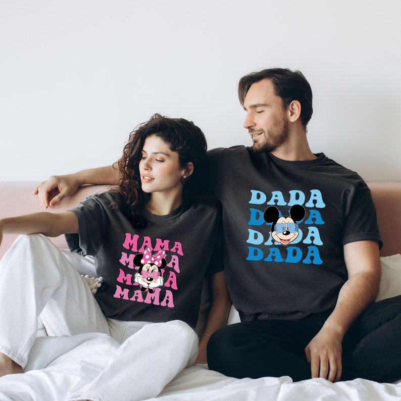 May include: A couple wearing matching black t-shirts with a graphic of Mickey Mouse. The woman's shirt says "MAMA" in pink letters with a pink bow on Mickey Mouse's head. The man's shirt says "DADA" in blue letters with blue sunglasses on Mickey Mouse's head.