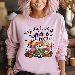 Op de afbeelding: Roze sweatshirt met een afbeelding van Mickey Mouse, Goofy en Winifred Sanderson uit Hocus Pocus. De tekst "It's just a bunch of Hocus Pocus" staat op het shirt gedrukt.