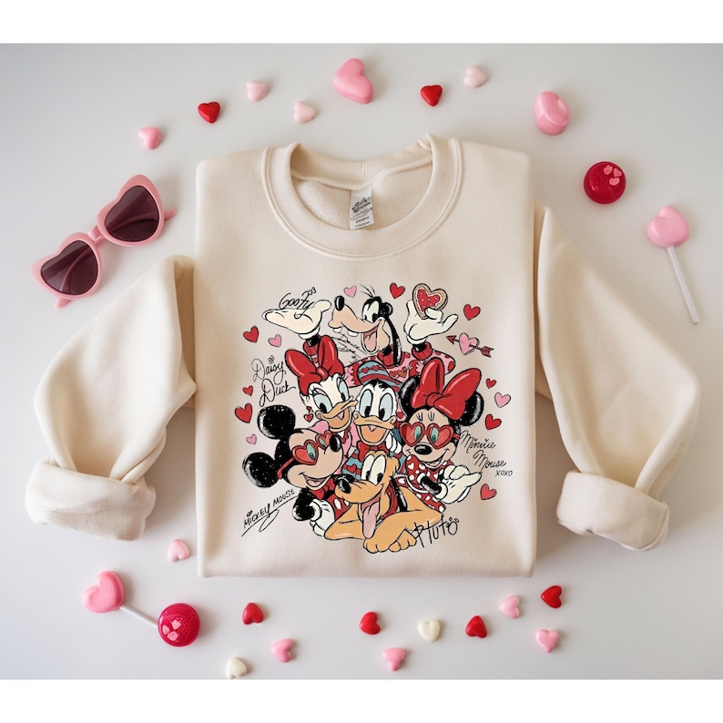 May include: Cream-colored sweatshirt featuring a graphic of Disney characters Mickey Mouse, Minnie Mouse, Donald Duck, Daisy Duck, Goofy, and Pluto, surrounded by hearts. The sweatshirt has long sleeves and is decorated with the text "Mickey Mouse" and "Minnie Mouse".