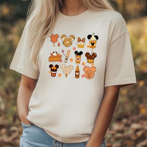 May include: Beige t-shirt featuring a collection of cartoon food items with Mickey Mouse ears. The illustrations include ice cream bars, pretzels, cupcakes, coffee, pumpkin pie, churros, cotton candy, and cookies. The design has a fall theme.