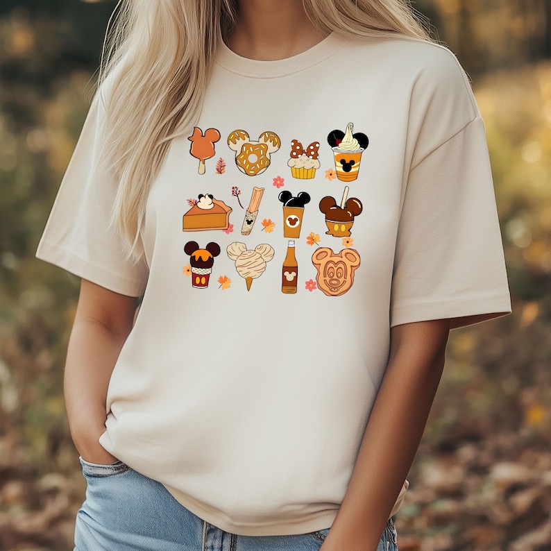 May include: Beige t-shirt featuring a collection of cartoon food items with Mickey Mouse ears. The illustrations include ice cream bars, pretzels, cupcakes, coffee, pumpkin pie, churros, cotton candy, and cookies. The design has a fall theme.