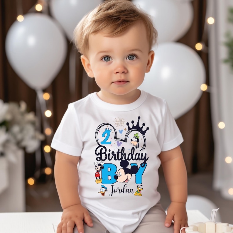 Op de afbeelding: Wit T-shirt met een blauw en zwart Mickey Mouse ontwerp. Het shirt heeft de tekst "2 Birthday Boy Jordan" met een kroon en vuurwerk.