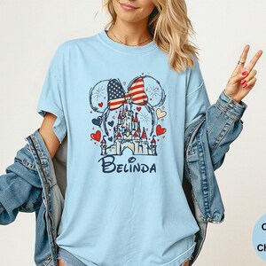May include: Light blue t-shirt featuring a graphic of a castle, Mickey Mouse ears with an American flag design, and the name "Belinda" in a script font. The design includes red and blue hearts. The shirt is a Comfort Colors Chambray.
