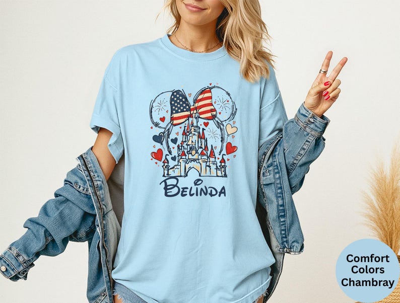 May include: Light blue t-shirt featuring a graphic of a castle, Mickey Mouse ears with an American flag design, and the name "Belinda" in a script font. The design includes red and blue hearts. The shirt is a Comfort Colors Chambray.